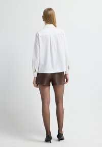 Witte blouse met lange mouwen en een kraag, met splitmanchetten, in combinatie met bruine leren shorts. Een paar simpele zwarte pumps maakt de look compleet.
