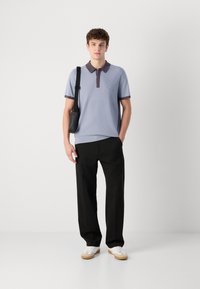 Camisa polo azul claro com tecido texturizado, gola escura em contraste e logotipo, combinada com calças pretas e ténis brancos.