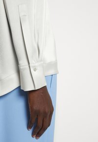 Light gray satin long-sleeve blouse cuff with button detail, paired with a light blue garment, showcasing a smooth texture and tailored fit.