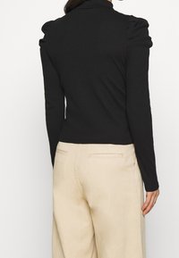 Top nero a maniche lunghe con spalle a sbuffo, realizzato in tessuto testurizzato. Indossato con pantaloni beige a vita alta con tasche posteriori.