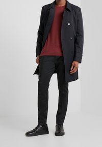 Trench-coat noir avec boutons double boutonnage, chemise bordeaux à col rond, pantalon noir slim et baskets noires, créant un look épuré.