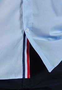 Chemise bleu clair à manches courtes avec boutonnage, présentant un panneau caché rayé rouge, blanc et marine le long de la couture latérale. Tissu lisse.