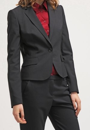 Vrouw in een op maat gemaakt zwart pak met een blazer met één knoop en bijpassende broek, gecombineerd met een donkerrood overhemd met knopen, staand tegen een effen achtergrond.