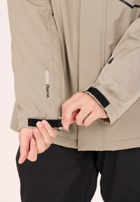 Beige vattentät jacka med justerbara kardborreband i ärmsluten. Funktioner inkluderar en slät yta, enkel design och synlig branding på ärmen.