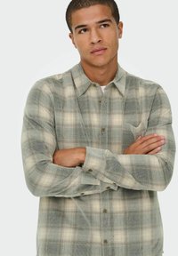 Zelena in bež karirana srajca, z gumbi in ovratnikom, dolge rokave in žep na prsih, izdelana iz teksturiranega blaga.