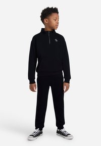 Gutt i svart zip-up hettegenser og joggebukser iført svarte og hvite sneakers, står og ser til siden mot en hvit bakgrunn.