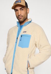 Ζακέτα Patagonia από φλις σε εκρού με ανοιχτομπλε λεπτομέρειες, φερμουάρ στην τσέπη στο στήθος, ψηλό γιακά, υφασμάτινη υφή και πλαϊνές τσέπες.