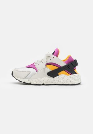 Biały, różowy i żółty sportowy sneaker z czarnym paskiem na pięcie i cętkowaną podeszwą, pokazany w profilu z prawej strony na białym tle.
