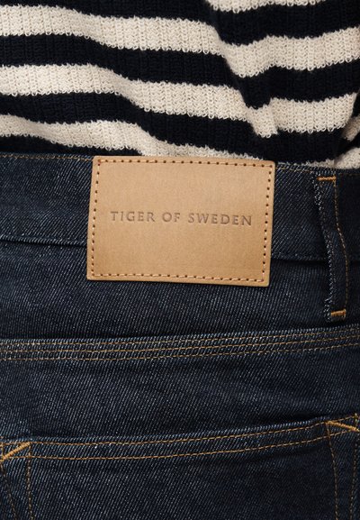 Pruhované černobílé tričko vsazené do tmavě modrých džínů s béžovou koženou nášivkou s nápisem "Tiger of Sweden" nad kapsou.