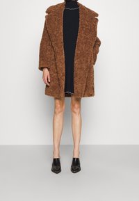 Brun, fluffig jacka med överdimensionerad krage; ihop med en ribbad svart klänning. Svarta, spetsiga skor fulländar outfiten.