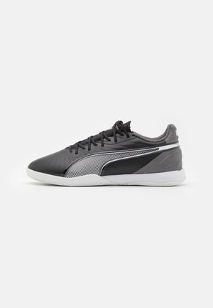 Chaussure de sport basse noire et grise avec lacets noirs, semelle blanche et logo blanc rayé sur le côté sur un fond blanc uni.