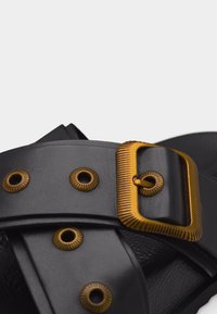 Μαύρο δερμάτινο λουράκι σανδάλου με χρυσή αγκράφα και ταιριαστά μεταλλικά τρύπες, εμφανίζοντας λεπτομερή υφή και ραφές.