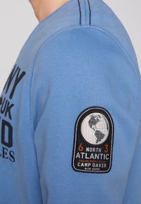 Hellblauer Pullover mit schwarzem Text und einem gestickten Aufnäher, der eine Weltkugel, Koordinaten und das Label "CAMP DAVID" am Ärmel zeigt.