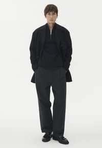 Cappotto nero sovrapposto a un pullover scuro, abbinato a pantaloni neri larghi e scarpe nere lucide. Insieme semplice e monocromatico.