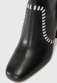 Bottines en cuir noir à bout carré, présentant des accents de couture blanche le long de la couture. Texture lisse avec une finition polie.