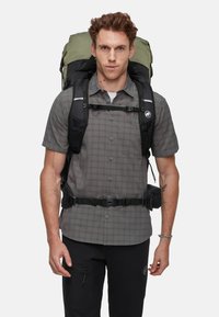Mochila con diseño gris y negro, correas ajustables y acentos metálicos. Llevada sobre una camisa de cuadros, con un bolsillo en los pantalones.
