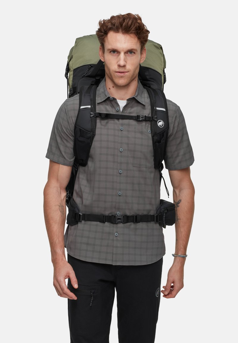 Mochila con diseño gris y negro, correas ajustables y acentos metálicos. Llevada sobre una camisa de cuadros, con un bolsillo en los pantalones.