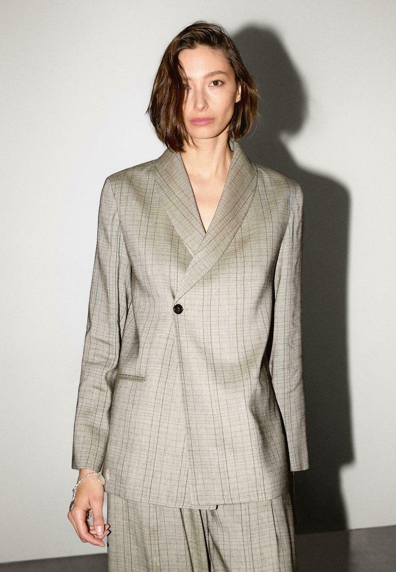 Lichtgrijze, geblokte blazer met een sjawl-kraag en enkele knoopsluiting. Textuurstof en getailleerde pasvorm met zijzakken.