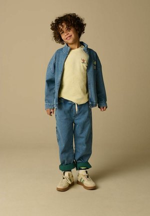 Enfant aux cheveux bouclés portant un sweat-shirt jaune, une veste en denim bleue et un pantalon, ainsi que des baskets blanches, debout sur un fond beige uni.