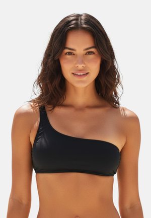 Donna con capelli mossi e scuri che indossa un top bikini nero monospalla, sorridente e rivolta verso avanti su uno sfondo chiaro uniforme.