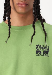 Sweatshirt din bumbac verde cu guler rotund, având un logo grafic negru și text pe pieptul stâng. Materialul este neted și are o croială lejeră.