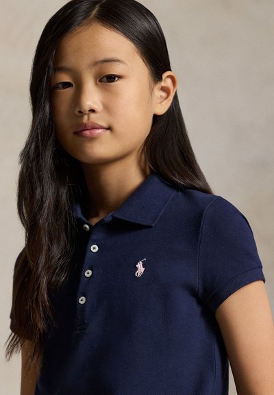 Jauna mergina su ilgais tamsiais plaukais, vilkinti tamsiai mėlyną polo marškinėlį su rožine išsiuvinėta logotipu, stovi prieš neutralią foną.