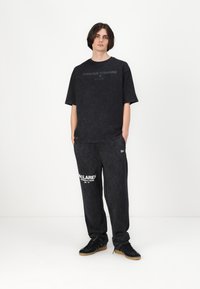 Melnā pārāk liela T-krekls ar uzrakstu "MŪŽĪGI UZ PRIEKŠU" tumšā drukā, saskaņot ar sporta biksēm, kuru priekšā ir "MCLAREN FORMULA 1 TEAM" logo baltā krāsā.