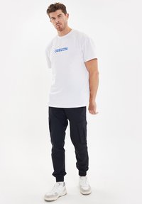 Witte t-shirt met korte mouwen met blauwe "OREGON" tekst, gecombineerd met zwarte cargobroek en witte sneakers met grijze accenten. Duidelijke details zichtbaar.