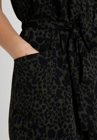 Tutina con stampa leopardata in verde scuro e nero, caratterizzata da maniche corte, un nastro in vita e ampie tasche laterali per un tocco di funzionalità.