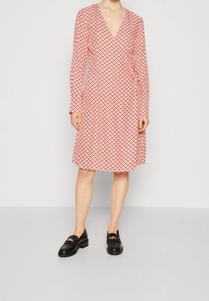 Kobieta ubrana w sukienkę o długości do kolan, z długim rękawem, zdobioną czerwonym wzorem w kształcie muszli, oraz czarne mokasyny z ozdobą ze złotą klamrą, stojąca na szarej podłodze.