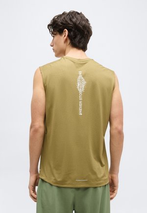 Homme aux cheveux foncés portant un débardeur vert olive avec un texte vertical blanc "Never Stop Playing" et un motif d'empreinte de chaussure au dos.