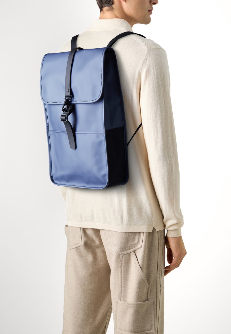 Zaino blu a forma rettangolare, realizzato in materiale sintetico liscio, con chiusura a pattina e una tracolla nera. Presenta tasche laterali e cuciture a contrasto.