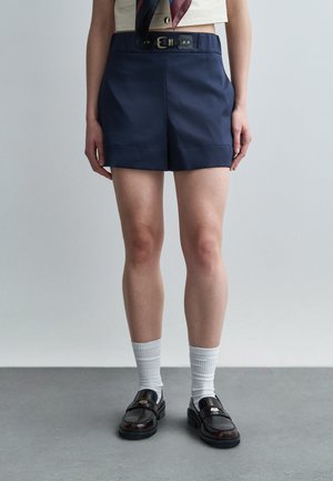 Osoba stojaca na sivom podlahe pri hladkej stene, oblečená v námornícky modrých šortkách s opaskom, bielych ponožkách a tmavohnedých mokasínach.