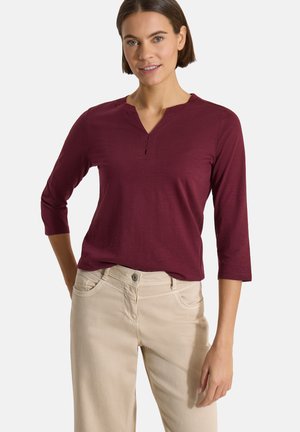 Femme aux cheveux courts et bruns, portant un haut bordeaux à manches longues avec un petit col en V et un pantalon beige, debout avec une main dans la poche.