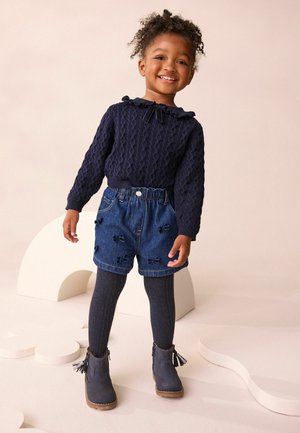 Marineblaue Strickpullover mit Rüschensaum, kombiniert mit Jeansshorts mit Schleifenakzenten. Dunkle Strumpfhosen und graue Stiefeletten runden das Outfit ab.