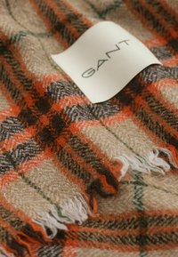 Écharpe en laine à carreaux avec des rayures beige, orange, marron et vert, bords frangés, et une étiquette blanche avec l'inscription "GANT."
