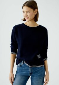 Navyblauer Pullover aus weichem Material mit Rundhalsausschnitt, gestreiften Bündchen und einem kleinen Logopatch an der unteren Seite. Die Jeans sind hellblau.