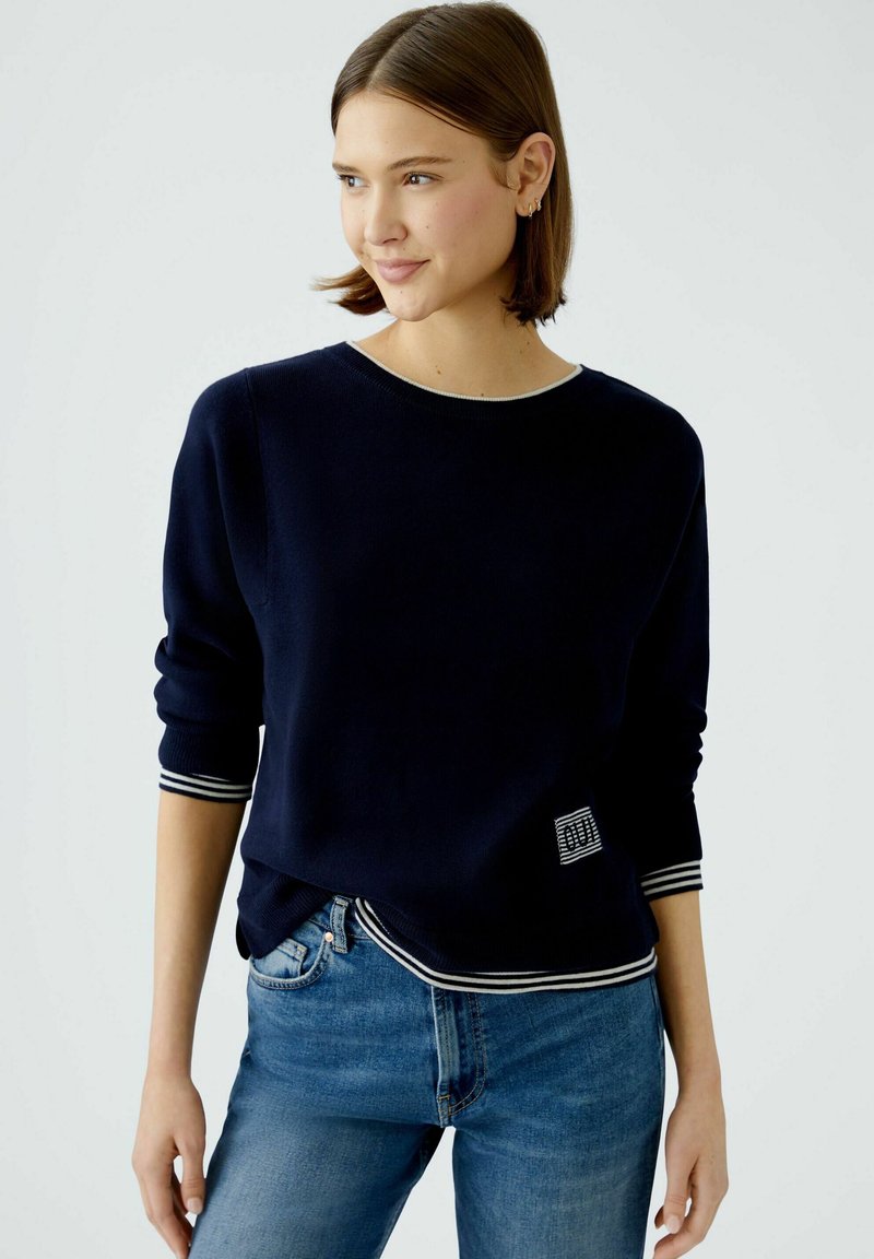 Navyblauer Pullover aus weichem Material mit Rundhalsausschnitt, gestreiften Bündchen und einem kleinen Logopatch an der unteren Seite. Die Jeans sind hellblau.