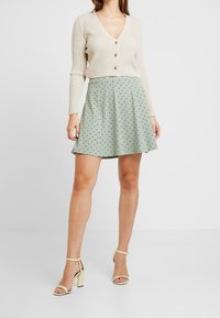 Beige geribbelde cardigan met knopen, gecombineerd met een groene schommelrok met stippen. Model draagt gouden blokhakken.