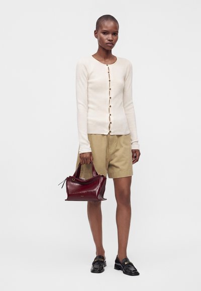 Femme portant un haut beige boutonné à manches longues, un short marron clair, des mocassins noirs, tenant un sac à main en cuir bordeaux sur un fond clair uni.