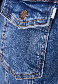 Denim fabric pocket detail featuring a bronze button, orange stitching, and a textured surface with a blue color gradient.