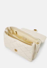 Sac à main crème matelassé avec une fermeture à bouton-pression doré. Comprend une poche intérieure zippée et une texture lisse. Forme rectangulaire compacte.