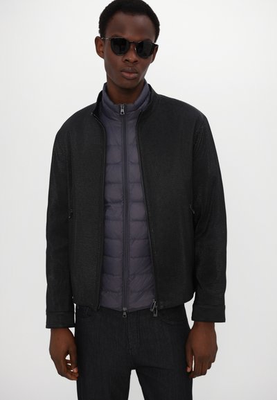 Schwarze perforierte Jacke über einer dunkelgrauen Steppinnenschicht, mit hohem Kragen und Reißverschlusstaschen. Getragen mit dunklen Jeans und Sonnenbrille.