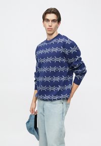 Μπλε σκούνο πλεκτό πουλόβερ με λευκό μοτίβο από συρματόπλεγμα, στρογγυλή λαιμόκοψη και ριμπ μανσέτες. Το μοντέλο κρατά μπλε καπέλο.