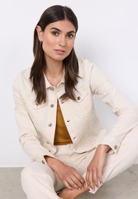 Giacca di jeans beige chiaro con bottoni metallici, caratterizzata da due tasche sul petto e un fit rilassato, abbinata a un top senape e pantaloni beige.