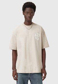 Young man with short curly hair wearing a beige oversized T-shirt with white graphic text and blue jeans, standing against a plain background.