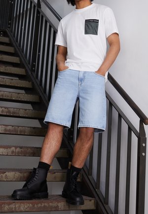 Homme portant un t-shirt blanc avec une poche noire, un short en jean bleu clair, des chaussettes noires et des bottes noires, debout sur un escalier industriel en métal.
