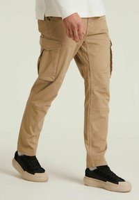 Khaki spodnie cargo z wieloma kieszeniami, zwężanymi nogawkami i gładką fakturą, w połączeniu z czarnymi niskimi trampkami z kremową podeszwą.