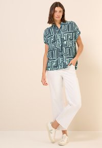 Koszulka z krótkim rękawem w kolorze turkusowym z czarnymi wzorami geometrycznymi, zestawiona z białymi szerokimi spodniami oraz beżowymi trampkami z zielonymi akcentami.