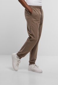 Lichtbruine sweatpants met een elastische tailleband, zijzakken en elastische boorden, met een logo op het linkerpoot. Witte sneakers worden gedragen.